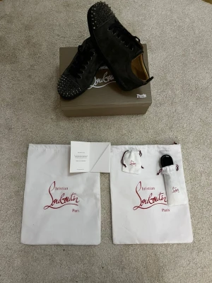 Christian Louboutin grå sneakers med nitar - Säljer ett par grå sneakers från Christian Louboutin i mocka med coola silverfärgade nitar på tån. Skorna har rund tå, snörning och platt sula. Innersulan är beige med guldlogga. Medföljer originalkartong, snöre, spikar och dustbags. Skorna har storlek 41 och pris kan självklart diskuteras!