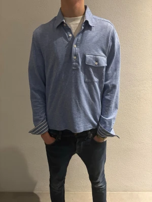 Blå skjorta från BOSS med knappar - Snygg blå henley från BOSS med vitmelerad ton och klassisk krage. Skjortan har en bröstficka med knapp, vita knappar framtill och randiga detaljer på insidan av ärmsluten. Tillverkad i mjuk bomull som känns chill och skön mot huden. Riktigt riktigt fet, hade velat behålla om den var lite längre i storleken (jag är 193).