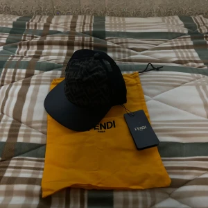 Svart Fendi keps med mesh och logga - Snygg svart keps från Fendi med klassiskt FF-mönster på framsidan och mesh på sidorna för extra ventilation. Justerbar rem med guldfärgad Fendi-logga bak. Tillverkad i polyester och bomull. Perfekt för dig som vill ha en exklusiv streetstyle-accessoar. Storlek 58cm (S) 