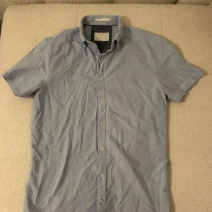 Ljusblå kortärmad skjorta Tailored Originals - Snygg ljusblå skjorta från Tailored Originals med korta ärmar och klassisk krage. Skjortan har knappar hela vägen framtill och är tillverkad i ett mjukt bomullstyg. 