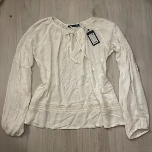 Vit blus med knyt och spetsdetalj - Supersöt vit blus från lager 157 med långa ärmar, knytning vid halsen och fina spetsdetaljer nedtill. Blusen har en luftig känsla och lätt struktur i tyget, perfekt för en fräsch och stilren look.