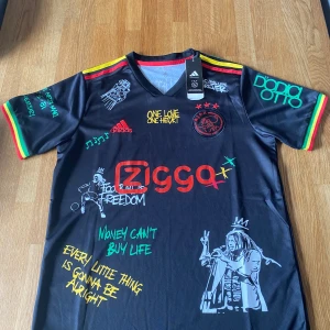 Ajax x Bob Marley Adidas fotbollströja M - Unik Ajax fotbollströja från Adidas med Bob Marley-tema. Svart fotbollströja med färgglada detaljer i rött, gult och grönt på ärmarna. Tryck och broderier med texter, motiv och Ajax-logga. Coola grafiska prints och Ziggo sponsor på bröstet. Aeroready-material för skön känsla. Passar även i S. Buda gärna😁