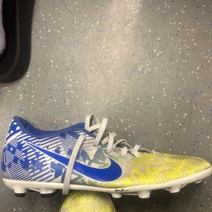 Neymars skor - Snygga fotbollsskor från Nike med blå och gula grafiska mönster, vit bas och blå detaljer. Skorna har snörning och en klassisk rund tå. Perfekta för dig som vill sticka ut på planen med stilrena färger och cool design.