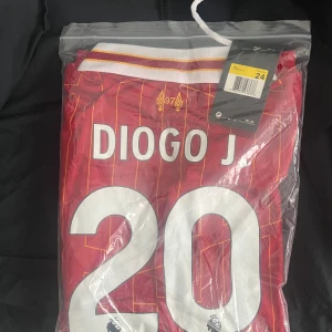 Liverpool Diogo Jota röd fotbollströja - Snygg röd Liverpool fotbollströja med Diogo Jota och nummer 20 på ryggen. Tröjan har tunna gula ränder och vit krage med röda och gula detaljer. Officiell Premier League-tryck och klubbmärke. Perfekt för dig som vill visa ditt stöd på matchen.