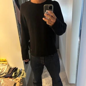 Svart stickad tröja från Louis Vuitton - Svart stickad tröja från Louis Vuitton med diskret LV-logga i metall vid nederkanten. Tröjan har rund hals, ribbade detaljer vid axlar och muddar samt långärmad passform. Materialet är mjukt och ger en clean och stilren look.