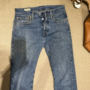 Levi's 501 blå jeans W28 L32 - Min brors gamla Klassiska blå Levi's 501 jeans med raka ben och knappgylf. Jeansen har fem fickor, tydliga kontrastsömmar och den ikoniska läderpatchen bak i midjan. Tillverkade i slitstark bomullsdenim och har den klassiska röda Levi's-taggen på bakfickan.