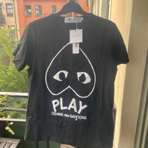 Svart t-shirt Comme des Garçons Play - Svart t-shirt från Comme des Garçons Play med stort vitt hjärta med ögon och logga på bröstet. Klassisk rund hals och korta ärmar. Skön bomullskvalitet och stilren design som sticker ut med den ikoniska grafiken framtill.