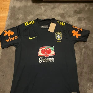 Brasilien Nike fotbollströja svart - Snygg svart Brasilien fotbollströja från Nike med gula och orangea detaljer. Tryck med CBF-emblem, sponsorer och Guaraná Antarctica-logga på bröstet. Kortärmad modell i lätt och ventilerande material, perfekt för träning eller match.