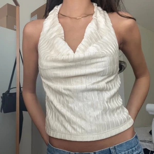 Vit halterneck topp med struktur - Säljer en snygg vit halterneck topp med draperad urringning och strukturerat tyg. Toppen är ärmlös och har öppen rygg, perfekt för varma dagar. Materialet känns lätt och har en subtil glans. Passar dig som gillar trendiga och stilrena plagg.