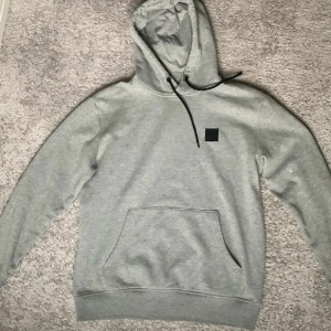 Grå hoodie med svart patch - Snygg grå hoodie med svart patch på bröstet och svarta snören i huvan. Klassisk känguruficka framtill och ribbade muddar vid ärmslut och nederkant. Perfekt för en chill och avslappnad stil. Bra skick.