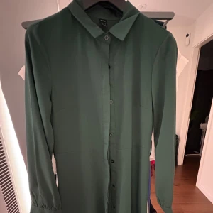 Grön skjortklänning från Weekday - Stilren grön skjortklänning från Weekday med knappar hela vägen framtill och klassisk krage. Långärmad modell i mjukt tyg, perfekt för dig som gillar minimalistisk stil.