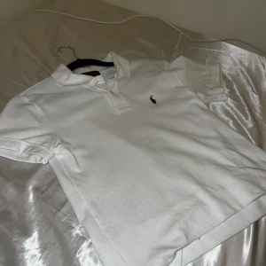 Vit Polo Ralph Lauren Piké  - Vit klassisk t-shirt från Polo Ralph Lauren i storlek M. T-shirten har korta ärmar, krage och två knappar framtill. Den är gjord i mjuk bomull och har den ikoniska broderade loggan på bröstet. Perfekt för en clean och stilren look. OBS Tröjan är i junior storlek M