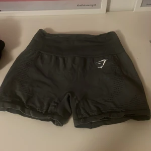 Mörkgrå Gymshark träningsshorts - Snygga mörkgrå träningsshorts från Gymshark med hög midja och ribbad linning. Materialet är stretchigt syntet och har ventilerande detaljer på sidorna. Perfekta för gymmet eller löpning, med diskret logga framtill.