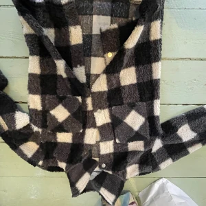 Rutig teddy-overshirt från Kappahl - Mysig och oversized teddy-overshirt från Kappahl i svart och vit rutig design. Skjortan har stora bröstfickor, tryckknappar och lång ärm. Perfekt för lager-på-lager och ger en cool, avslappnad vibe. Den är verkligen så snygg och perfekt nu till hösten! Säljer då den är liite för kort i ärmarna på mig