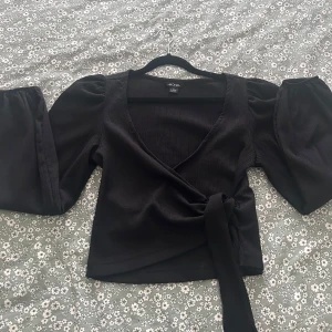 Svart omlottopp med puffärm från Monki - Svart omlottopp från Monki med långa puffärmar och knytband i midjan. Toppen har v-ringning och är tillverkad i ett ribbat, mjukt material som ger en snygg struktur. Perfekt för dig som gillar trendiga detaljer och en feminin siluett.