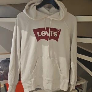 Vit hoodie från Levi's med röd logga - Säljer en vit hoodie från Levi's i storlek S. Tröjan har en stor röd Levi's-logga på bröstet, huva med snörning och en klassisk känguruficka framtill. Perfekt för dig som gillar en enkel och stilren streetlook.
