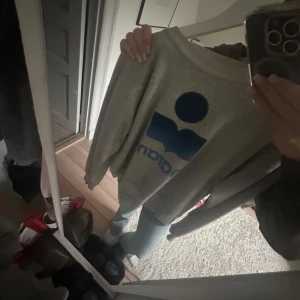  Isabel Marant tröja - Säljer en grå sweatshirt från Isabel Marant med stort blått logotryck framtill. Tröjan har en klassisk rund halsringning och långa ärmar. Perfekt för en chill och stilren look, funkar lika bra till jeans som till mjukisbyxor.