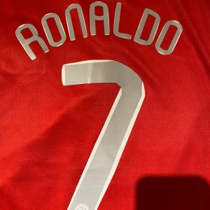 Manchester United Ronaldo 7 Finaltröja - Säljer en ikonisk röd Manchester United fotbollströja från Champions League-finalen 2008 med Ronaldo och nummer 7 på ryggen. Tröjan har broderat klubbmärke, AIG-logga, NikeFIT Dry-material och coola detaljer som 'The Red Devils'-patch. Perfekt för dig som älskar fotbollshistoria.