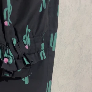 Svarta byxor med kaktusmönster - Unika svarta byxor med grönt och rosa kaktusmönster. Byxorna har snörning vid bensluten för en cool detalj och är tillverkade i ett mjukt, lätt tyg som känns skönt mot huden. Perfekta för dig som vill sticka ut med en lekfull stil.