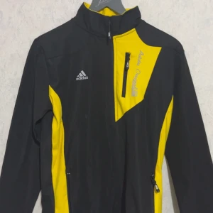 Svart och gul vindjacka från Adidas - Sportig vindjacka från Adidas i svart och gul med cool asymmetrisk design. Jackan har hög krage, dragkedjor fram och på fickor, samt Adidas-logga på bröstet. Perfekt för dig som gillar att sticka ut och vill ha en jacka med snygga detaljer.