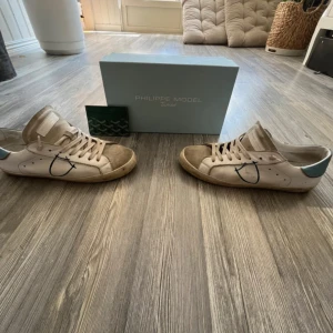Philippe Model vita sneakers strl 40 - Snygga vita Philippe Model sneakers med beige mockadetaljer på tån och plös, blå hälparti med röd kant och klassisk logga. Skorna har snörning, rund tå och platt gummisula. Unik blå detalj på sidan och stilren design som sticker ut.