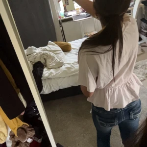 Ljusrosa blus med volang - Supersöt ljusrosa blus med korta, vida ärmar och smock i midjan som ger en snygg volang nertill. Perfekt att styla med jeans för en chill men ändå söt look. Blusen är i mjuk bomull och har en avslappnad passform.