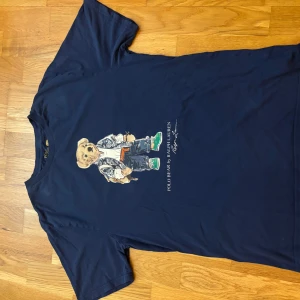 Blå Polo Bear t-shirt från Ralph Lauren - Mörkblå t-shirt från Ralph Lauren med ikoniskt Polo Bear-tryck på bröstet. Klassisk rund hals och korta ärmar. Tillverkad i mjuk bomull som är skön mot huden. Perfekt för dig som vill ha en cool och tidlös look med en twist. Den är XL i barnstorlek, 170