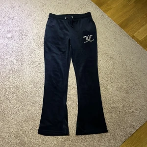 Juicy Couture barn mörk blå velourbyxor - Mörk blå velourbyxor från Juicy Couture med bootcut-snitt och glittrande JC-logo i strass på ena höften. Obs! Barnstorlek - 134/140! Byxorna har snörning i midjan och två bakfickor. Perfekta för en chill och trendig look.