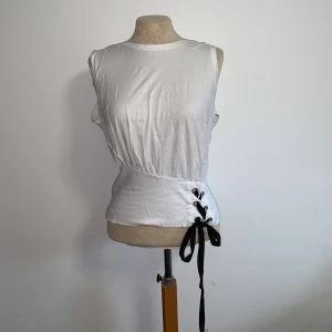 Vit ärmlös topp med snörning från Zara - Säljer en vit ärmlös topp från Zara med cool snörningsdetalj i svart på sidan. Perfekt för en chill och stilren look. Passformen är normal och toppen är enkel att matcha till olika outfits.