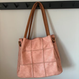Rosa axelväska med bruna handtag - Säljer en stor rosa axelväska med bruna handtag och dekorativa sömmar i rutmönster. Väskan har dragkedjor på sidorna och ett rymligt innerfack med flera mindre fack. Perfekt för dig som vill ha plats för allt du behöver under dagen. Den är köpt i Spanien.💓💓