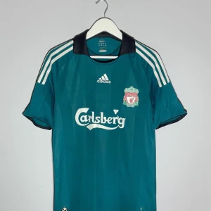 Liverpool Gerrard Adidas tröja - Snygg Liverpool fotbollströja från Adidas med Steven Gerrard och nummer 8 på ryggen. Tröjan är mörkgrön med svarta detaljer och vita ränder på axlarna. Klassisk Carlsberg-logga på bröstet och Liverpool FC-emblem. Tillverkad i lätt polyester, perfekt för match eller träning.