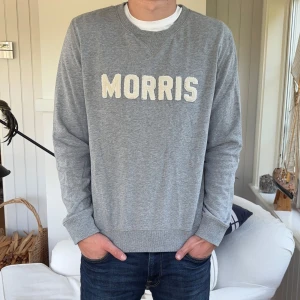Grå sweatshirt från Morris - Säljer en grå sweatshirt från Morris med vit broderad logga framtill. Tröjan har rund hals, ribbade muddar och är långärmad. Perfekt för chill dagar och enkel att matcha med jeans eller joggers.