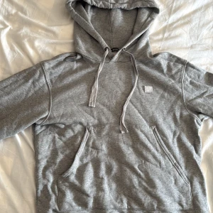 Grå hoodie från Acne Studios - Snygg grå hoodie från Acne Studios med klassisk face patch på bröstet. Tröjan har huva med snörning, magficka och är tillverkad i mjuk bomullsmix. Perfekt för chill dagar och streetstyle vibes.