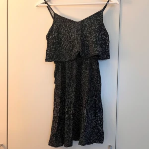 Svart lång prickig klänning - Svart långklänning med vita prickar och smala axelband. Klänningen har ett lager över bysten med en liten öppning framtill och ett mjukt, flowigt fall. Perfekt för dig som gillar en chill och stilren look med lite extra detaljer. Materialet känns lätt och luftigt. Total längd: ca 80cm (går att justera med axelbanden). 