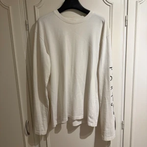 Vit långärmad Kenzo tröja - Säljer en stilren vit långärmad tröja från Kenzo med svart logotyptryck på ena ärmen. Gjord i mjuk bomull, perfekt för en clean och modern look. Passar dig som gillar minimalistisk design och streetwear.