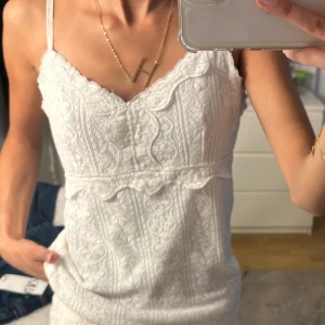 Vit spetsig linnetopp med smala band - Supersöt vit linnetopp med smala axelband och v-ringning. Toppen har ett detaljerat spetsmönster och en liten volangkant under bysten. Perfekt för en fräsch och romantisk look, lätt att matcha med jeans eller kjol.