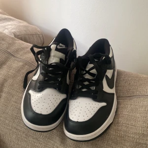 Nike Dunk Low svart/vit sneakers - Säljer ett par Nike Dunk Low sneakers i svart och vitt. Klassisk siluett med svart Swoosh, perforerad tå och vit bas. Skorna har snörning och platt sula, tillverkade i skinn och syntet. Perfekta för dig som gillar streetstyle och vill ha en ikonisk look.