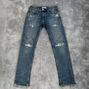 Replay jeans blå distressed 30/32 - Snygga blå jeans från Replay med slitna detaljer och råa benslut. Klassisk femficksmodell med raka ben och coola slitningar på knäna och framtill. Jeansen är i bomull och har en avslappnad vibe, perfekt för dig som gillar en lite ruffare stil. Hål över knät.