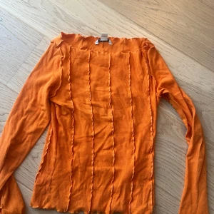 Orange långärmad topp från Monki - Säljer en orange långärmad topp från Monki i ribbat material. Toppen har dekorativa vågiga sömmar längs hela plagget och en rak, något vid halsringning. Sitter tight och har en modern, trendig look som sticker ut.