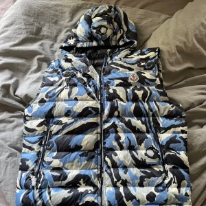 Moncler dunväst med blå camomönster - Säljer en fet dunväst från Moncler med blått, svart och vitt camomönster. Västen har huva, dragkedja framtill och två fickor. Perfekt för dig som vill sticka ut och hålla värmen. Moncler-logga på bröstet och cool streetkänsla.