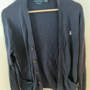 Mörkblå kofta från Polo Ralph Lauren - Säljer en klassisk mörkblå kofta från Polo Ralph Lauren i mjuk pima bomull. Koftan har knappar framtill, långa ärmar och den ikoniska broderade loggan på bröstet. Perfekt att slänga över en t-shirt eller skjorta för en chill och stilren look.