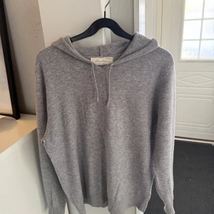 Grå kashmirhoodie från Sayless - Mjuk och lyxig grå hoodie från Sayless i kashmir. Klassisk modell med huva och dragsko, samt långa ärmar och ribbade muddar. Perfekt för dig som gillar en clean och stilren look med premiumkänsla.