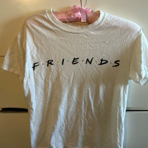 Vit FRIENDS t-shirt från Primark - Vit t-shirt från Primark med FRIENDS-tryck på bröstet i svart text och färgglada prickar mellan bokstäverna. Klassisk rund hals och korta ärmar. Tillverkad i 100% bomull, perfekt för dig som älskar tv-serien Friends.