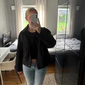 Svart kappa med knappar - Säljer en svart, kort kappa med klassisk krage och stora fickor framtill. Jackan har tre knappar och detaljer vid ärmsluten. Materialet är tjockt och känns som ull, perfekt för kyliga dagar. Snygg och stilren modell som är enkel att matcha.