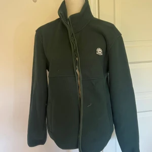 Mörkgrön fleecejacka från Reserved - Mörkgrön fleecejacka från Reserved med hög krage och dragkedja framtill. Jackan har en broderad patch på bröstet med texten 'Reach the Peak 1969 1992'. Perfekt för kyliga dagar och enkel att slänga på sig över en hoodie.