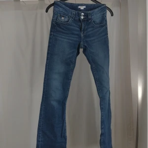 Blå bootcut jeans med dubbla knappar - Snygga blå jeans med bootcut passform och dubbla knappar i midjan. Klassisk femficksmodell med lätt slitning och raka ben som får en liten utsvängning nertill. Perfekta för dig som gillar en tidlös men trendig look. Från Gina youth stl 164