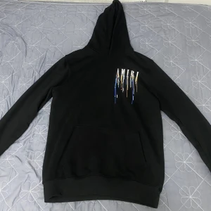 Svart AMIRI hoodie med färgstänk - Svart hoodie från AMIRI med stor huva och känguruficka. Framtill och baktill finns AMIRI-logga med coolt färgstänksmönster i blått, vitt, gult och rött. Perfekt för dig som vill sticka ut med streetstyle. Mjuk och skön bomullskänsla.