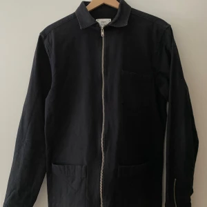 Svart overshirt med dragkedja - Stilren svart overshirt från Ciszere med rak passform, klassisk krage och hel dragkedja framtill. Jackan har tre fickor och är tillverkad i bomull. Perfekt för lager-på-lager och funkar till många olika outfits.