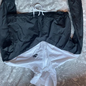 Nike svartvit vindjacka med huva - Nike vindjacka i svart och vitt med dragkedja framtill och huva. Jackan har en sportig look, snörning i midjan och klassisk Nike-logga på bröstet. Perfekt för dig som gillar streetstyle och vill ha något lätt och snyggt till vardags.