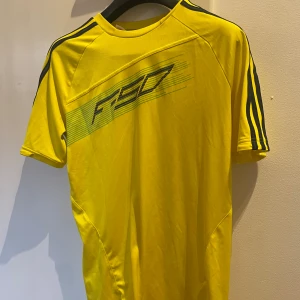 Gul sportig t-shirt med svarta ränder - Gul t-shirt från Adidas med svarta ränder på axlarna och tryck 'F50' på bröstet. Perfekt för träning eller sport, tillverkad i lätt polyester som andas. Klassisk rund hals och korta ärmar för en skön passform.